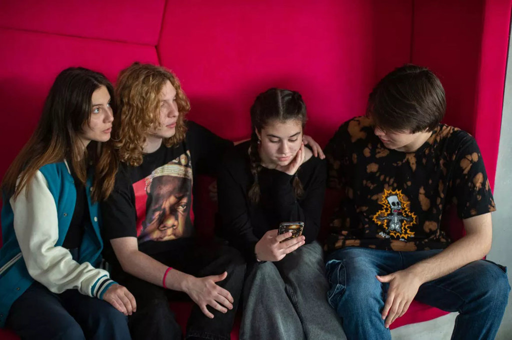 Quatro jovens adultos estão sentados em um sofá rosa vibrante, absortos em seus smartphones. A cena destaca o envolvimento dos jovens com a tecnologia digital e as redes sociais, refletindo temas de segurança na Internet, prevenção do cyberbullying e cidadania digital.