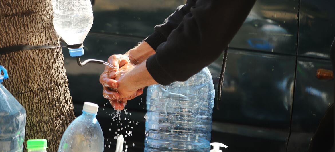 Um homem lava as mãos usando um dispensador de água improvisado criado a partir de uma garrafa de plástico presa a uma árvore, com várias outras garrafas grandes de água por perto. Esta cena retrata um esforço de ajuda humanitária para famílias deslocadas em Beirute, no Líbano.