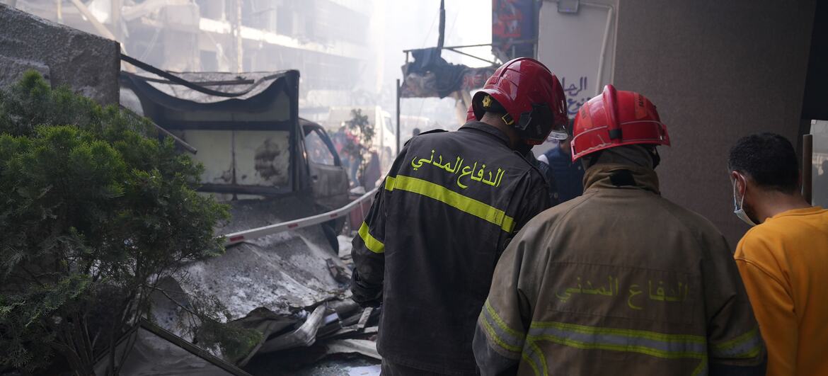 Dois bombeiros com capacetes vermelhos e uniformes com escrita árabe nas costas examinam uma cena de destruição com escombros e edifícios danificados em Beirute, no Líbano.
