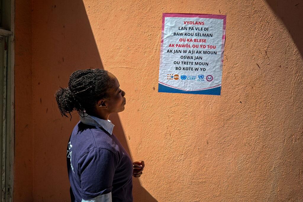 Uma mulher negra com cabelo trançado fica de perfil olhando para um pôster em uma parede laranja em Porto Príncipe, Haiti. O cartaz é uma campanha de sensibilização para a violência baseada no género, com logótipos de agências da ONU, incluindo UNOCHA, CERF e UNFPA.