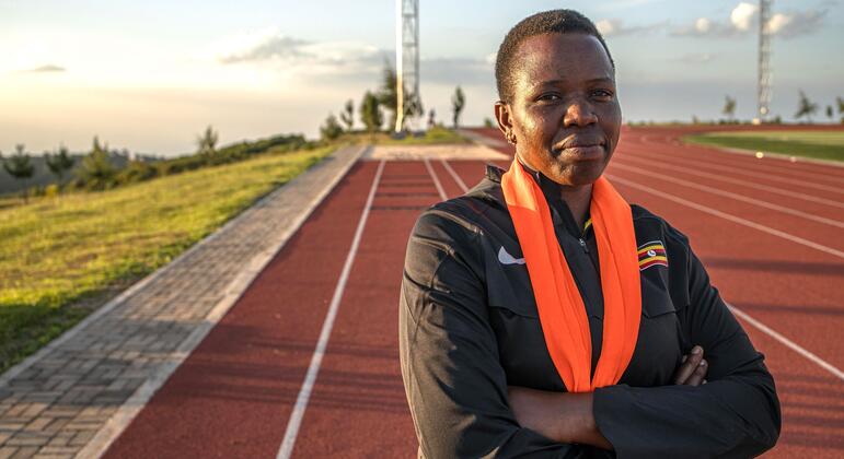 Retrato de uma treinadora no Centro Nacional de Treinamento em Alta Altitude Teryet, no distrito de Kapchorwa, Uganda. Ela está em uma pista de atletismo com os braços cruzados, vestindo uma jaqueta esportiva preta com um emblema da bandeira de Uganda e um lenço laranja.