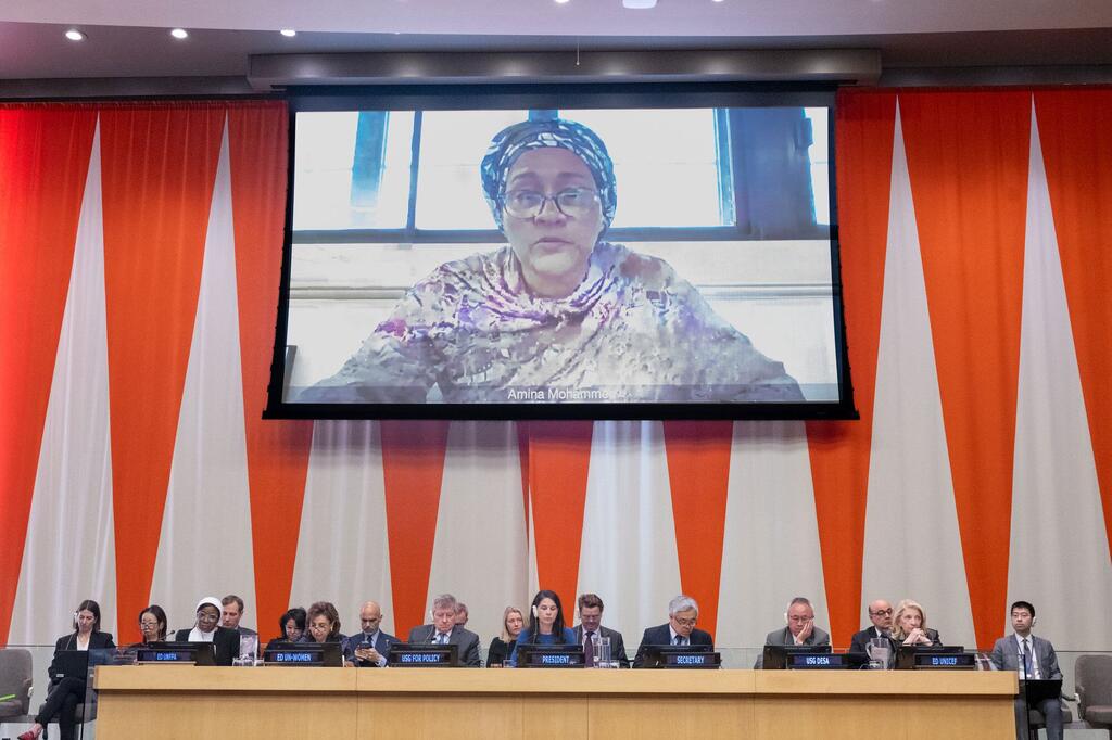 A vice-secretária-geral, Amina Mohammed, fala através de videoconferência em uma tela grande em uma reunião plenária da Assembleia Geral da ONU. Os delegados estão sentados em uma longa mesa em primeiro plano.